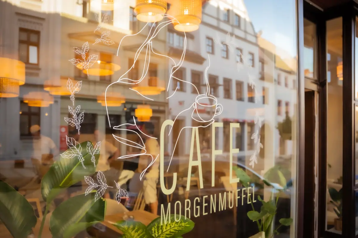 Schaufenster des Café Morgenmuffel mit Logo und leuchtenden Rattanlampen
